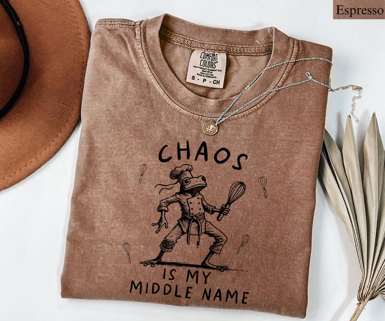 May include: Espresso-colored t-shirt with a crew neck and the text "CHAOS IS MY MIDDLE NAME." The graphic features a frog chef holding a whisk. A silver necklace with a pendant rests on the shirt.