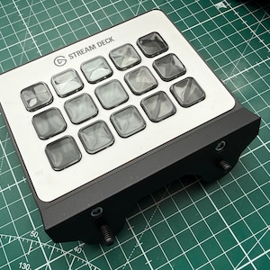 May include: A black and silver Stream Deck controller featuring a grid of fifteen square, translucent buttons on a metallic faceplate. The device has a black housing with two visible mounting screws at the front edge, designed for integration.