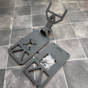 Op de afbeelding: Een matzwart metalen montagebeugelsysteem met een centrale verstelbare arm met een V-vormige houder, twee rechthoekige basisplaten met driehoekige uitsparingen, de tekst DRK. gegraveerd op de oppervlakken en een klein zakje met bevestigingsmateriaal.