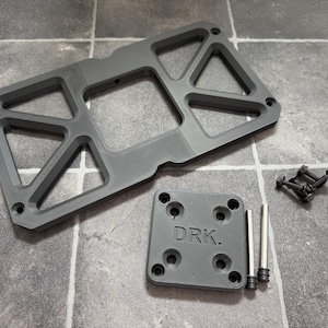 May include: A gray, geometric metal plate with a rectangular shape and cut-out sections. A smaller square plate with "DRK." text, screws, and metal rods are also present. The items are arranged on a tiled surface.