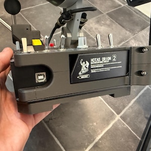 Op de afbeelding: Een grijze HOTAS ORION 2-controller met meerdere hendels, knoppen en een USB-poort. De controller heeft een zwart label met witte tekst en een logo. Het apparaat wordt in een hand gehouden.
