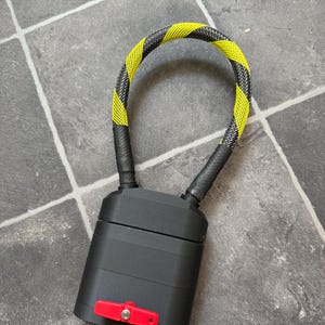 May include: A black rectangular lock body with a red lever switch on the front and a flexible, U-shaped cable loop extending from the top. The cable features a repeating diagonal pattern of bright yellow and black woven material.