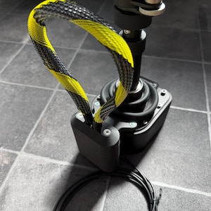 May include: A black joystick controller featuring a central vertical shaft with a rubberized base and a yellow and black braided cable loop attached to the side. The device includes a rectangular black housing with visible silver screws and a black cord.