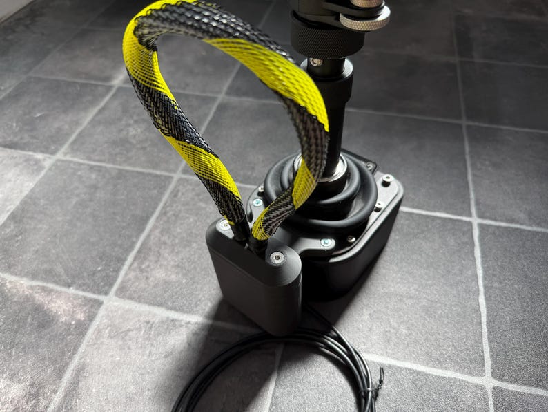 May include: A black joystick controller featuring a central vertical shaft with a rubberized base and a yellow and black braided cable loop attached to the side. The device includes a rectangular black housing with visible silver screws and a black cord.