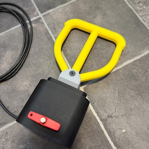 May include: A black rectangular device with a yellow D-shaped handle attached by a gray bracket. A small red rectangular plate with a silver circular button is mounted on the front of the black housing. A thin black cable extends from the side.