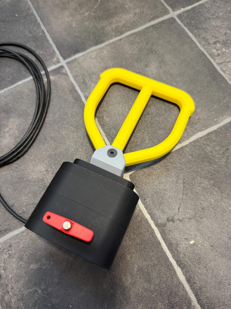 May include: A black rectangular device with a yellow D-shaped handle attached by a gray bracket. A small red rectangular plate with a silver circular button is mounted on the front of the black housing. A thin black cable extends from the side.