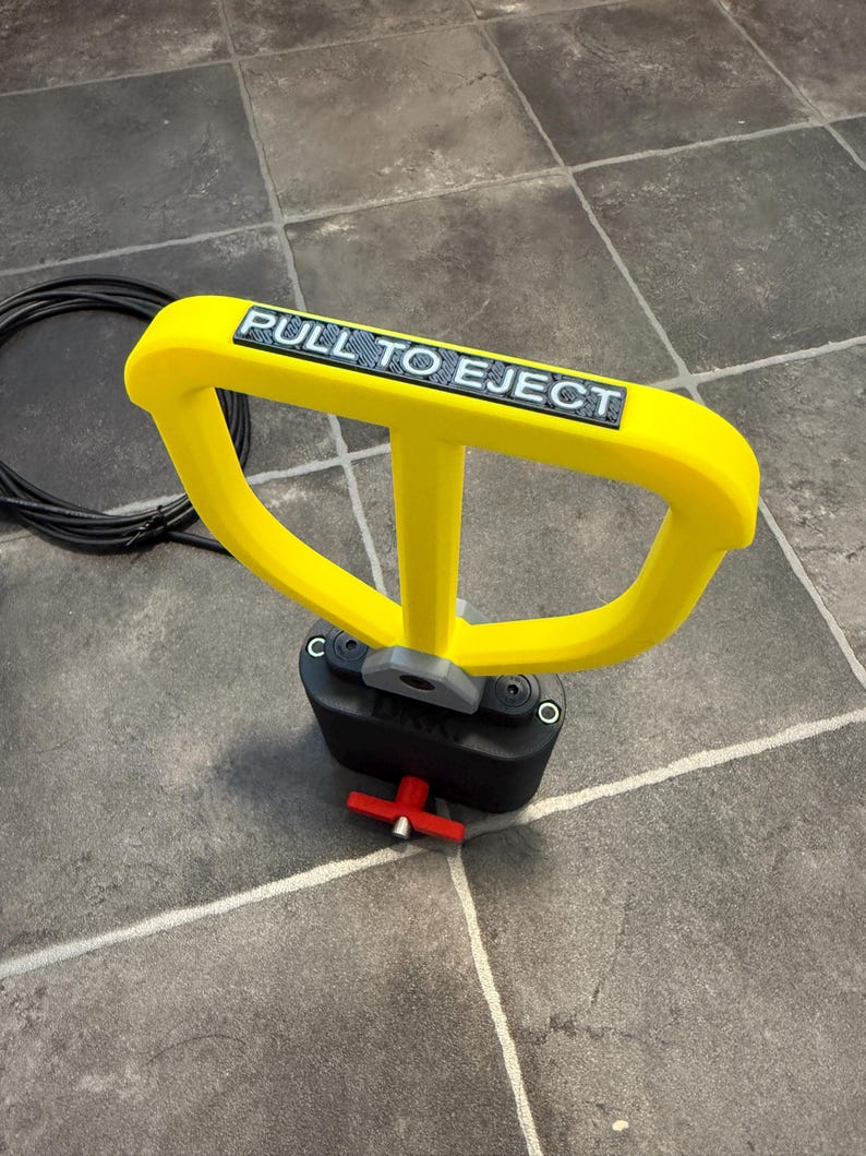 May include: A bright yellow handle with the text PULL TO EJECT in black letters, attached to a black base with a grey connector. A small red T-shaped lever is visible at the bottom of the black base, and a black coiled cable lies nearby.