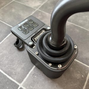 Könnte beinhalten: Ein schwarzer Joystick mit einem Bedienfeld mit mehreren quadratischen Tasten. Der Joystick hat einen gebogenen Griff und eine Basis mit silberner Zierleiste. Das Produkt ist für Spiele oder Flugsimulationen konzipiert.