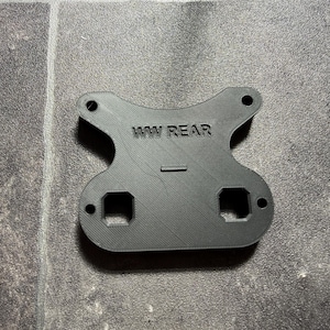 May include: A black, flat, 3D-printed component with a symmetrical, curved shape. It features the embossed text 'WW REAR' in the center, two circular holes at the top, two square cutouts at the bottom, and two smaller circular holes on the sides.
