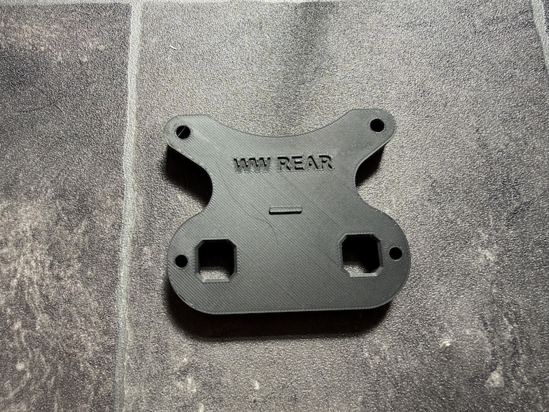 May include: A black, flat, 3D-printed component with a symmetrical, curved shape. It features the embossed text 'WW REAR' in the center, two circular holes at the top, two square cutouts at the bottom, and two smaller circular holes on the sides.