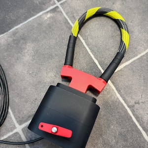 May include: A black and red security lock featuring a flexible cable with a yellow and black hazard pattern. The device includes a rectangular black housing with a red lever and a metallic silver pin, connected to a U-shaped locking mechanism.