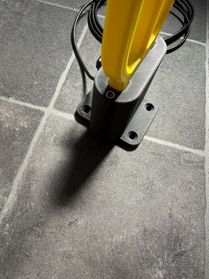 May include: A black 3D-printed mounting base with four circular screw holes, featuring a vertical yellow handle component inserted into the top. A thin black cable extends from the rear of the base, which has a textured, layered surface finish.
