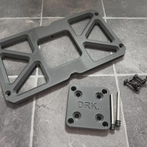 May include: A matte gray rectangular mounting bracket with triangular cutouts, a smaller square plate embossed with the text DRK., two silver metal pins, and four small black screws. The components are arranged together on a flat surface.
