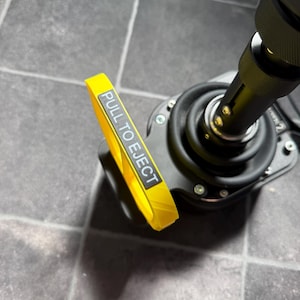 May include: A black mechanical joystick base featuring a prominent yellow pull-tab handle. The handle is rectangular with a grey label that clearly displays the white text PULL TO EJECT. The device includes a black rubber boot and metal hardware.