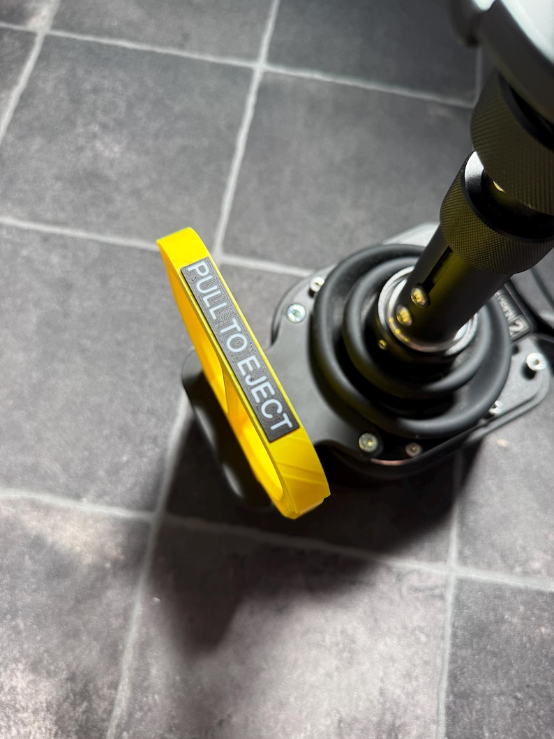 May include: A black mechanical joystick base featuring a prominent yellow pull-tab handle. The handle is rectangular with a grey label that clearly displays the white text PULL TO EJECT. The device includes a black rubber boot and metal hardware.