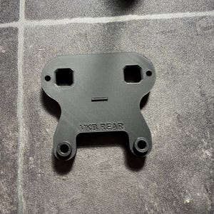 May include: A black, flat, 3D-printed mounting bracket with a symmetrical shape. The piece features two square cutouts at the top, two circular mounting holes, and two raised circular attachment points at the base. The text VKB REAR is embossed.