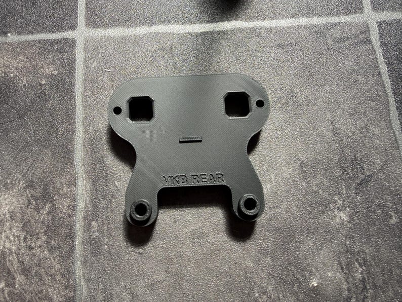 May include: A black, flat, 3D-printed mounting bracket with a symmetrical shape. The piece features two square cutouts at the top, two circular mounting holes, and two raised circular attachment points at the base. The text VKB REAR is embossed.