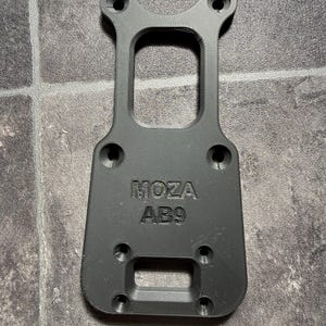May include: A black 3D-printed mounting plate featuring the embossed text MOZA AB9. The component has a rectangular base with rounded corners, a central cutout, and six countersunk screw holes arranged for attachment to a flat surface.