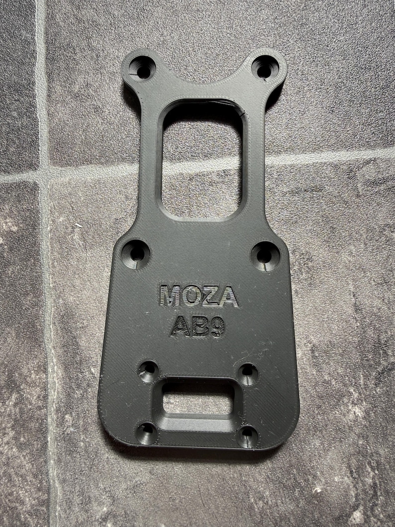May include: A black 3D-printed mounting plate featuring the embossed text MOZA AB9. The component has a rectangular base with rounded corners, a central cutout, and six countersunk screw holes arranged for attachment to a flat surface.