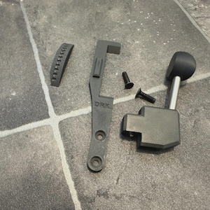 May include: An assortment of gray mechanical parts, including a lever with a black knob, a curved piece with small holes, a bracket with the letters "DRK", and two screws. The parts are arranged on a gray tiled surface.
