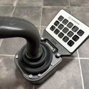 Op de afbeelding: Een zwarte en zilveren Stream Deck-controller met een gebogen handvat. De controller heeft een wit paneel met 15 zwarte vierkante knoppen en de tekst "STREAM DECK". De basis heeft de tekst "DRK.  X-HAPSODON 2".