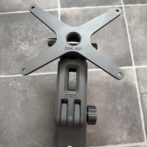 Könnte beinhalten: Ein schwarzer, verstellbarer Monitorhalter mit kreuzförmiger Oberseite und dem Text "DRK. ICP". Die Halterung hat eine vertikale Stütze mit Einstellknöpfen und dem Text "DRK.".