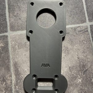 May include: A matte black, 3D-printed mounting plate with a rectangular top section featuring four circular screw holes and a large central circular cutout. The lower section has four smaller screw holes and a rectangular cutout, with the text AVA embossed.