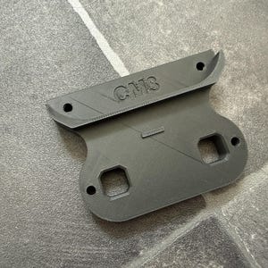 May include: A dark gray 3D printed mounting bracket featuring the embossed text CM3. The component has a wide top edge with two circular holes, a central body with two square cutouts, and two additional circular holes on the lower section.