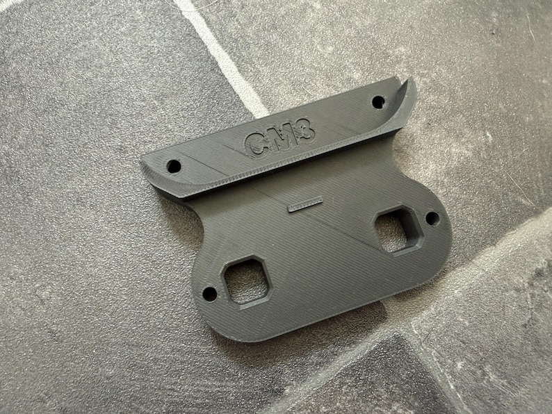 May include: A dark gray 3D printed mounting bracket featuring the embossed text CM3. The component has a wide top edge with two circular holes, a central body with two square cutouts, and two additional circular holes on the lower section.