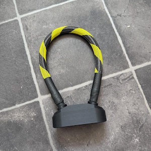 Könnte beinhalten: Ein U-förmiges Fahrradschloss mit einem flexiblen Kabel, das mit einem schwarzen und neongelben geflochtenen Netzmuster überzogen ist. Die Basis des Schlosses ist ein massives, mattschwarzes rechteckiges Gehäuse, das die beiden Kabelenden verbindet.