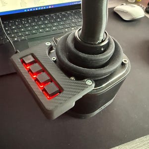Puede incluir: Un joystick negro con un panel de fibra de carbono con cuatro botones rojos iluminados. El joystick tiene una bota de goma negra y está conectado a un portátil. Un ratón blanco y rosa es visible en el fondo.