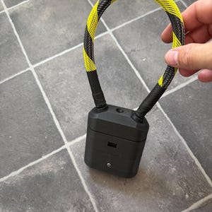 May include: A black rectangular charging case for wireless earbuds featuring a flexible, braided cable loop with a yellow and black diagonal hazard stripe pattern attached to the top, including a small circular button and a charging port on the front.