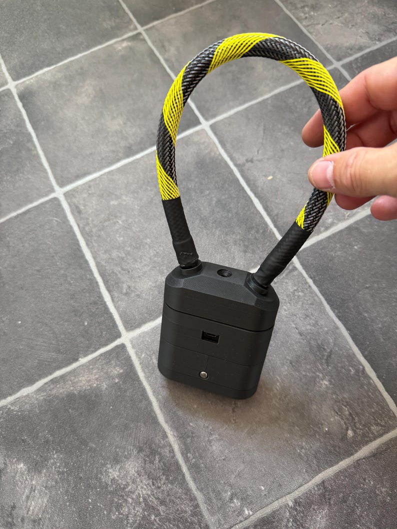 May include: A black rectangular charging case for wireless earbuds featuring a flexible, braided cable loop with a yellow and black diagonal hazard stripe pattern attached to the top, including a small circular button and a charging port on the front.