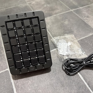 Op de afbeelding: Een zwart rechthoekig apparaat met 20 vierkante knoppen, aan de boven- en zijkanten gemarkeerd met "DRK". Een zwarte USB-kabel en een kleine zak met accessoires liggen naast het apparaat. De items liggen op een grijze tegelvloer.