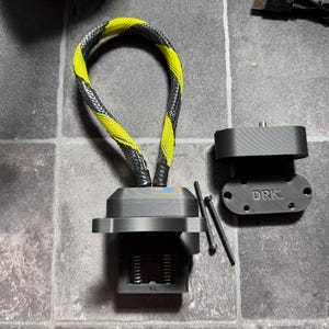 May include: A black 3D-printed computer cooling component featuring a yellow and black braided cable loop, a mounting bracket with the text DRK. embossed on the surface, and two thin metal pins for assembly, all presented as a set of hardware parts.