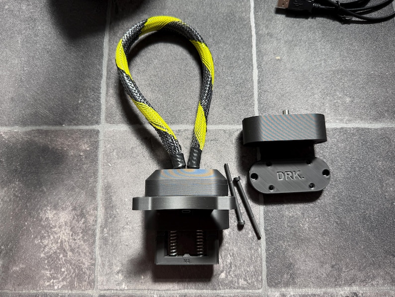 May include: A black 3D-printed computer cooling component featuring a yellow and black braided cable loop, a mounting bracket with the text DRK. embossed on the surface, and two thin metal pins for assembly, all presented as a set of hardware parts.