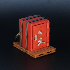 May include: A miniature red safe with a silver handle and dial, secured with gray straps. It rests on a brown pallet base and includes a keychain ring. The safe is a novelty item.