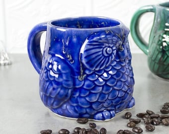 May include: Two ceramic owl mugs, one blue and one green, with a scattering of coffee beans in front of them.
