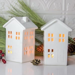 May include: Two white ceramic houses with cut-out windows and a lit candle inside. The houses are sitting on a white surface with pine branches and pine cones.