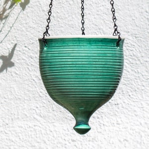 以下が含まれることがあります： リブ模様と尖った底を持つ緑色のセラミック製の吊り下げプランター。プランターは黒いチェーンで吊り下げられています。