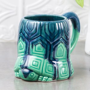 Puede incluir: Una taza de cerámica verde y azul con forma de tortuga. La taza tiene un asa y un diseño detallado de caparazón de tortuga.