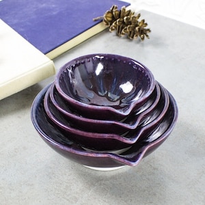 May include: Set of four nesting bowls in shades of purple and blue with a white glaze. The bowls are made of ceramic and have a unique, hand-painted design.