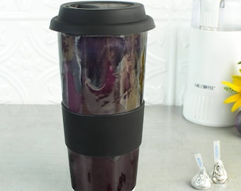 May include: A black and purple ceramic travel mug with a black silicone lid and a black silicone band around the middle. The mug is sitting on a gray surface with coffee beans scattered around it.