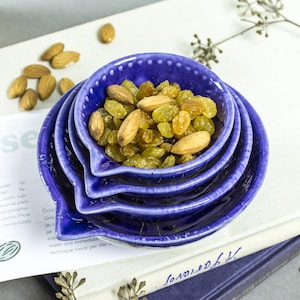 May include: Four blue ceramic nesting bowls with a white speckled glaze. The top bowl is filled with golden raisins and almonds.
