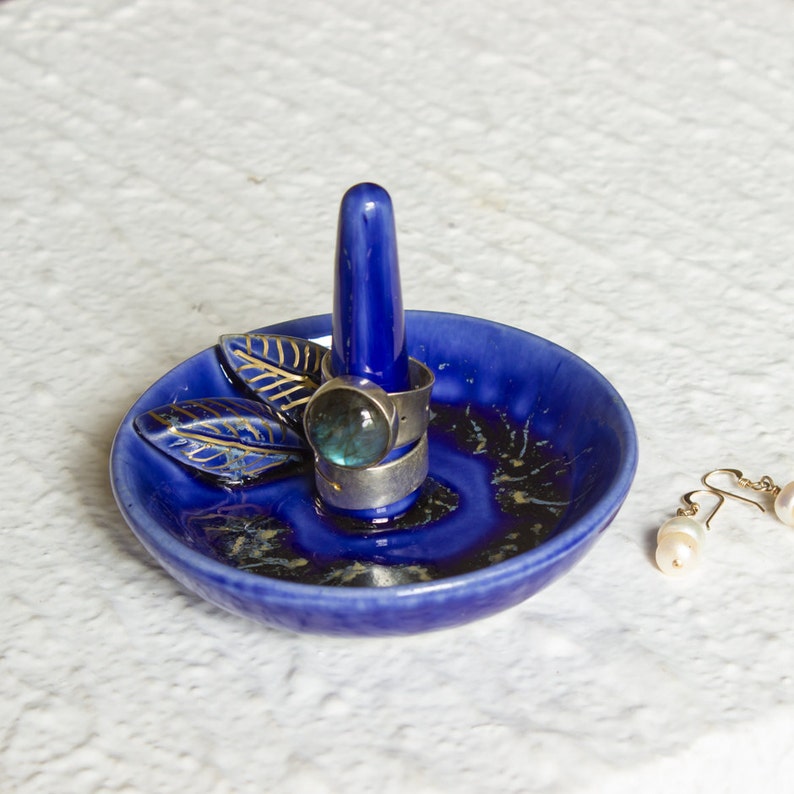 May include: A cobalt blue ceramic ring holder with a central post and a small dish. The dish features gold leaf accents and holds two silver rings with a large, round, blue-green stone. A pair of pearl earrings rests nearby.