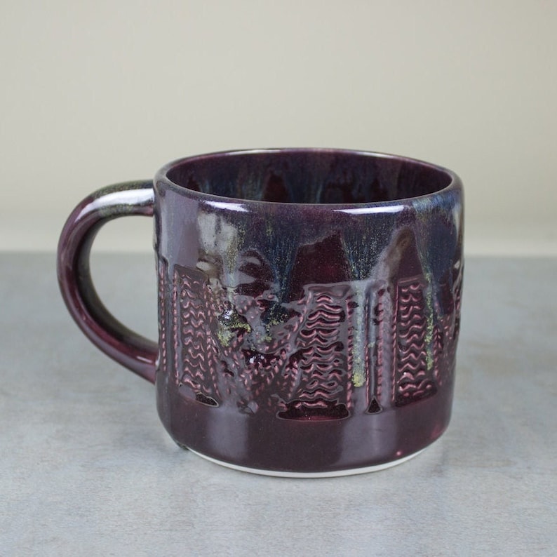 May include: A handmade ceramic mug with a dark purple glaze. The mug features a textured design on the exterior and a curved handle. The interior has a dark blue and green glaze. The mug is suitable for hot or cold beverages.