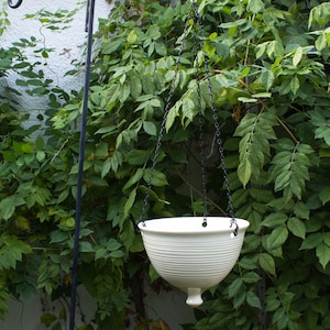 May include: White ceramic hanging planter with a ribbed design. The planter is suspended from a black chain and hangs from a metal hook.