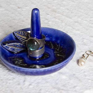 May include: A cobalt blue ceramic ring holder with a central post and a small dish. Two silver rings with a large, round, blue-green stone are on the post. A pair of pearl earrings rests beside the dish.