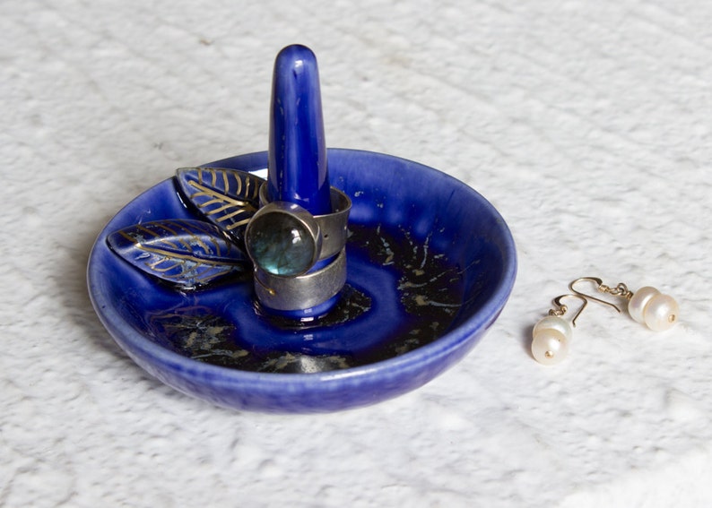 May include: A cobalt blue ceramic ring holder with a central post and a small dish. Two silver rings with a large, round, blue-green stone are on the post. A pair of pearl earrings rests beside the dish.
