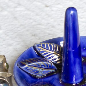 May include: A cobalt blue ceramic ring holder with a tall, rounded post and a shallow dish base. The dish features two leaf-shaped accents with gold detailing. The ring holder is designed to display rings.